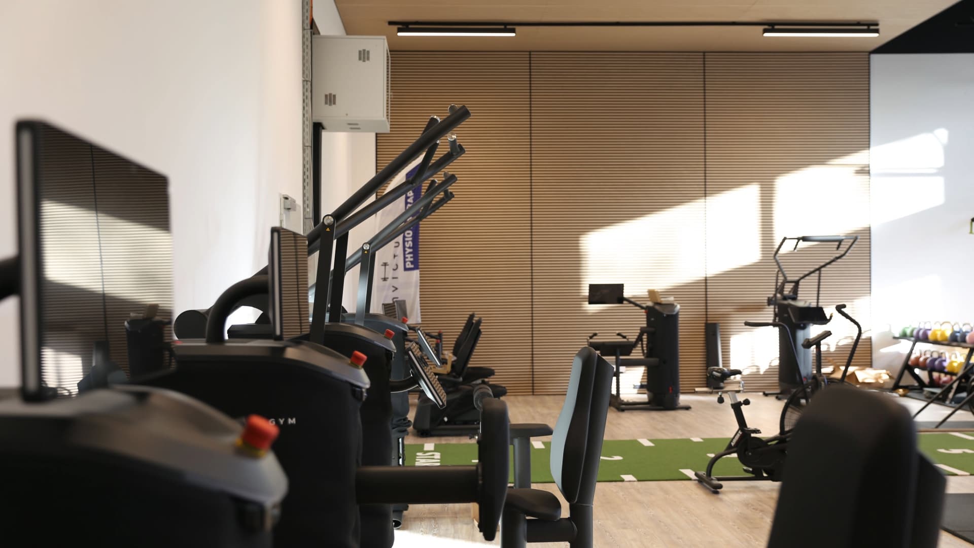 Blick über die Trainingsfläche des Invictus Sports Club: vorne die EGYM-Geräte, dahinter die grüne Sprintbahn, ein Airbike und Kettlebells vor der Holzlamellenwand.